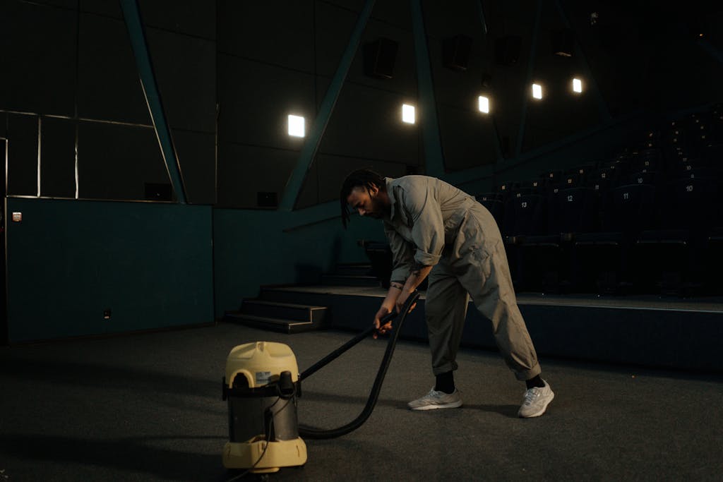 A cinema employee cleaning the theater floor with a vacuum cleaner under dim lights.